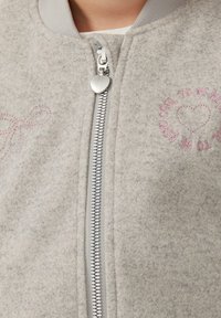 Giubbotto bomber in feltro grigio chiaro con zip metallizzata e tirazip a forma di cuore. Presenta dettagli in rosa ricamati e un design a fiocco.