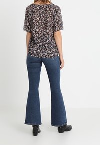 Hedvábná blůza s květinovým vzorem a krátkými rukávy, tmavé pozadí, a přiléhavé tmavě modré rozšiřující se džíny. Černé kozačky dokončují celkový vzhled.