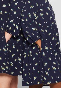 Navy sukienka w kwiatowy wzór, z bufkami, warstwowym krojem i kieszeniami po bokach. Materiał wydaje się lekki i zwiewny.