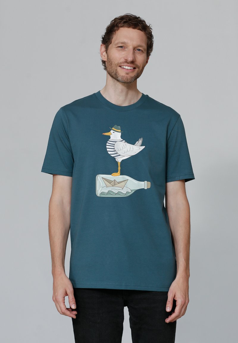 Teal katoenen T-shirt met een cartoonzeemeeuw in een gestreept shirt en hoed, staande op een fles met een papieren bootje.