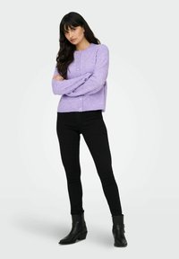 Pull en tricot lilas avec un col rond et un détail de boutons, associé à un jean skinny noir et des bottines noires. Texture lisse, coupe décontractée.