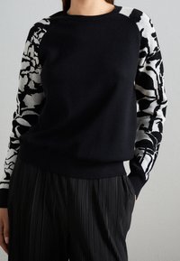 Svart tröja med mönstrade grå och vita ärmar. Mjuk stickad kvalitet, rundad halslinning, ribbade muddar och nederkant, kombinerad med plisserade svarta byxor.