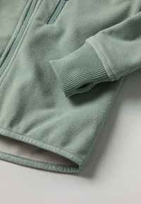 Fleece-takki vaaleanvihreänä, jossa on täysipitkä vetoketju, ribbireunukset ja logotiedot helmassa. Pehmeä rakenne ja kodikas ilme.