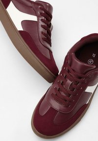 Burgunderfarbene Sneaker mit einem schlanken Design, aus Leder und Wildleder, weißen Akzenten an den Seiten, runden Schnürsenkeln und strukturierten braunen Sohlen.