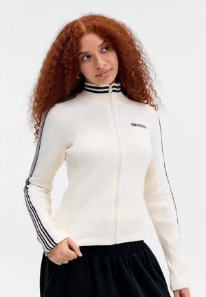 Žena s dlhými kučeravými červenými vlasmi, oblečená v bielu bundu na zips s čiernymi pruhmi na rukávoch a čiernych nohaviciach, pozerá sa do strany.