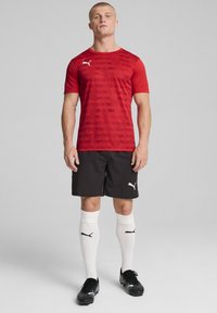 Camiseta deportiva roja con patrón texturizado, pantalones cortos negros, calcetines blancos con logo y zapatos de fútbol negros, aptos para actividades deportivas.