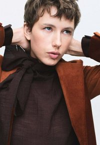 Blazer in suede marrone con tessuto scuro sotto, caratterizzato da un dettaglio di sciarpa annodata al colletto e motivi testurizzati sulla camicia.