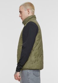 Olivgrüne wattierte Weste mit hohem Kragen, Reißverschluss und nahtlosem Design, getragen über einem schwarzen Langarmshirt.