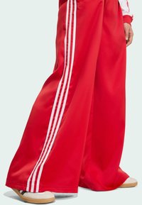 Red wide-leg pants made of smooth fabric, featuring three white side stripes. Paired with cream and brown footwear.