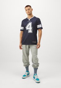 Tengeri focimez világos fehér részletekkel, 4-es számjeggyel. Szürke melegítőnadrág és teal, valamint lila részletekkel ellátott sneakers. Fehér háttér.