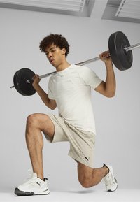 Camiseta atlética blanca con logo, pantalones cortos beige y barra de pesas negra. El calzado es blanco con detalles negros. Sujeto en posición de zancada.