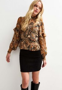 Długa bluzka z wzorem paisley z falbanami w kolorach pomarańczowym, czarnym i beżowym, zestawiona z czarną mini spódnicą z sztruksu i czarnymi kozakami za kolano.