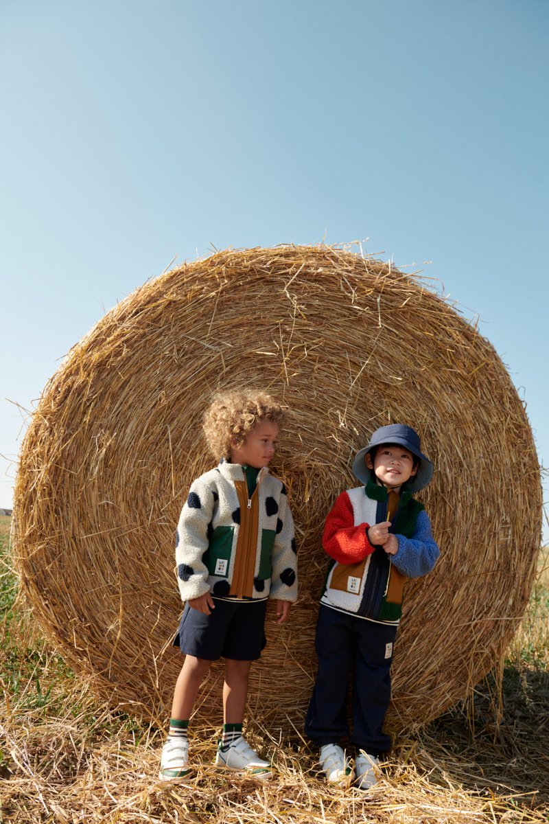 To børn står ved siden af en høstak. Den ene har en fleecejakke med polkaprikker og en grøn lomme; den anden har en fleecejakke i flere farver.
