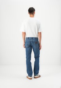 Blauwe spijkerbroek met een recht gesneden ontwerp, vijf zakken, zichtbare stiksels en een leren patch op de achterste tailleband. Witte t-shirt gedragen.