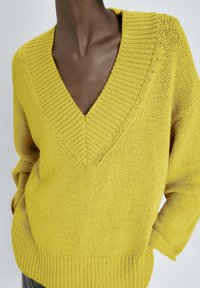 Pull jaune en maille avec un col en V profond, un col et un ourlet côtelés, présentant un motif texturé. Coupe ample avec épaules tombantes.