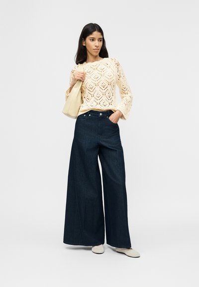 Νεαρή γυναίκα που φοράει μπεζ πλεκτό πουλόβερ και σκούρο τζιν με φαρδιά μπατζάκια, κρατώντας μπεζ τσάντα στον ώμο, στέκεται μπροστά σε απλό φόντο.