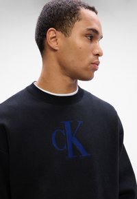 Čierna mikina s okrúhlym výstrihom a rebrovanými manžetami, s modrým logom "cK" na prednej strane. Materiál vyzerá štruktúrovaný a teplý.