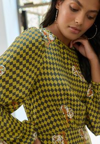 Zaļš pārbaudītais kleitas modelis ar ziedu akcentiem, ar garām piedurknēm un savilkta josta, izgatavots no gludas auduma ar drosmīgiem rakstiem.