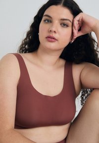 Brun, glatt stoff bralette med en bred, rund halslinje og tykke stropper. Den buede designen komplimenterer kroppens form, uten synlig maskinvare.