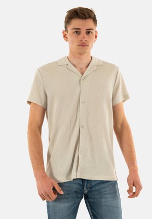 Chemise beige à manches courtes avec une surface texturée, fermeture par bouton et col, associée à un jean bleu.