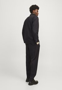 Completo formale nero con giacca e pantaloni su misura. Il tessuto appare liscio e con una leggera lucentezza. Presenta bottoni sui polsini e grandi tasche laterali.