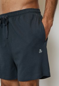 Mörkblå elastiska shorts med dragsko, sidofickor och en liten vit logotyp på vänster sida. Tillverkade av mjukt tyg.