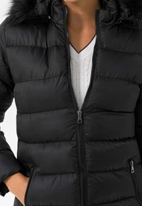 Sort pufferjakke med lynlås foran, der har en aftagelig hætte med pelsfor, quiltet tekstur og sidelommer. Hvid kabelstriktrøje under.