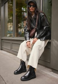 Botines de charol negro con suelas texturizadas, combinados con pantalones anchos beige y una chaqueta de piel negra. Los accesorios incluyen un bolso marrón.
