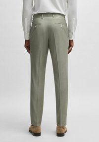 Pantaloni leggeri verdi chiari con gamba dritta, gamba affusolata, due tasche posteriori con bottoni, indossati con una camicia bianca a maniche lunghe e scarpe color nocciola.