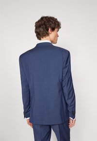 Navyblauer, taillierter Anzugjacke aus strukturiertem Stoff, mit einem Schlitz hinten, Knopfmanschetten und einem darunter sichtbar weißen Hemd.