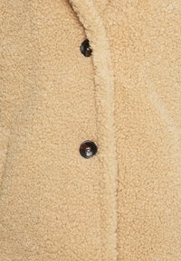 Miękki beżowy płaszcz o fakturalnym wykończeniu, z gładkim czarnym guzikiem i klasycznym kołnierzem.