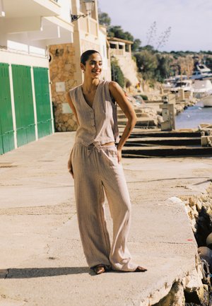 Kvinde iført en top med striber uden ærmer og bukser med vide ben, stående på en solbeskinnet promenade ved vandet med både og stenhuse i baggrunden.