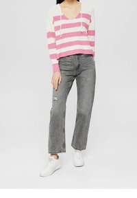 Persona vestida con un suéter a rayas rosas y blancas, jeans gris de cintura alta con un desgastado en la rodilla izquierda y zapatillas blancas, de pie sobre un fondo blanco.