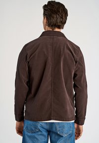 Chaqueta de algodón marrón con corte relajado, mangas largas y cuello, combinada con pantalones de mezclilla azules, vista desde la espalda.