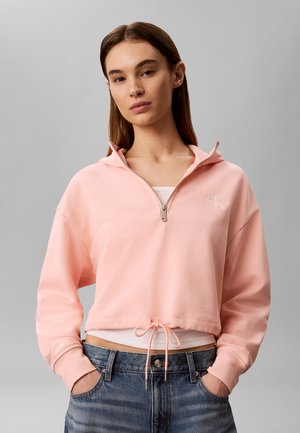 Jonge vrouw draagt een lichtroze cropped hoodie met halve rits en koord, wit ondershirt en blauwe spijkerbroek, staand tegen een grijze achtergrond.
