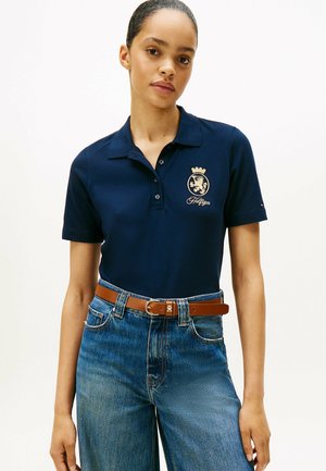 Polo bleu marine avec une finition texturée, arborant un emblème doré et un détail de couronne. Associé à un jean bleu taille haute et une ceinture marron.