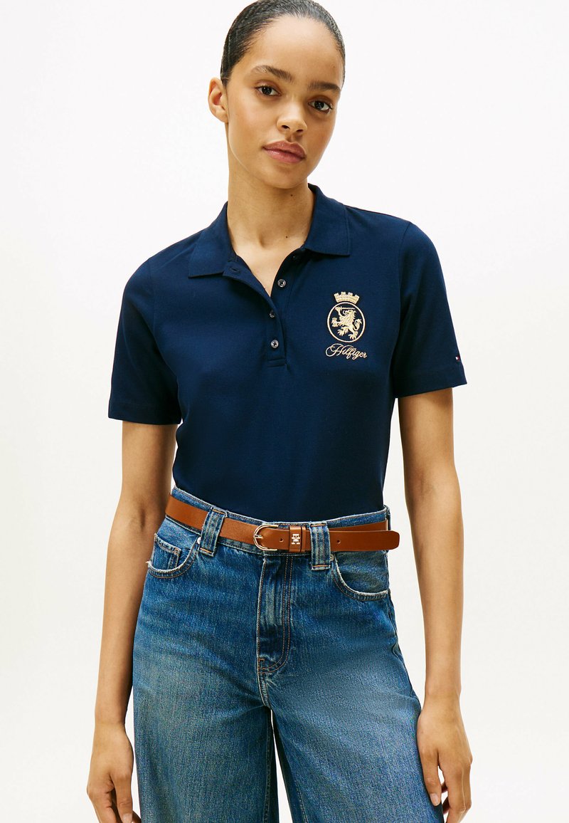 Granatowa koszulka polo o teksturowanym wykończeniu, z złotym emblematem i detalem korony. Połączona z niebieskimi dżinsami o wysokim stanie i brązowym paskiem.