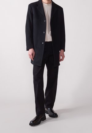 Manteau en laine noire avec col cranté, doté d'une fermeture à boutons. Associé à une chemise beige clair et un pantalon noir, complété par des bottes noires.