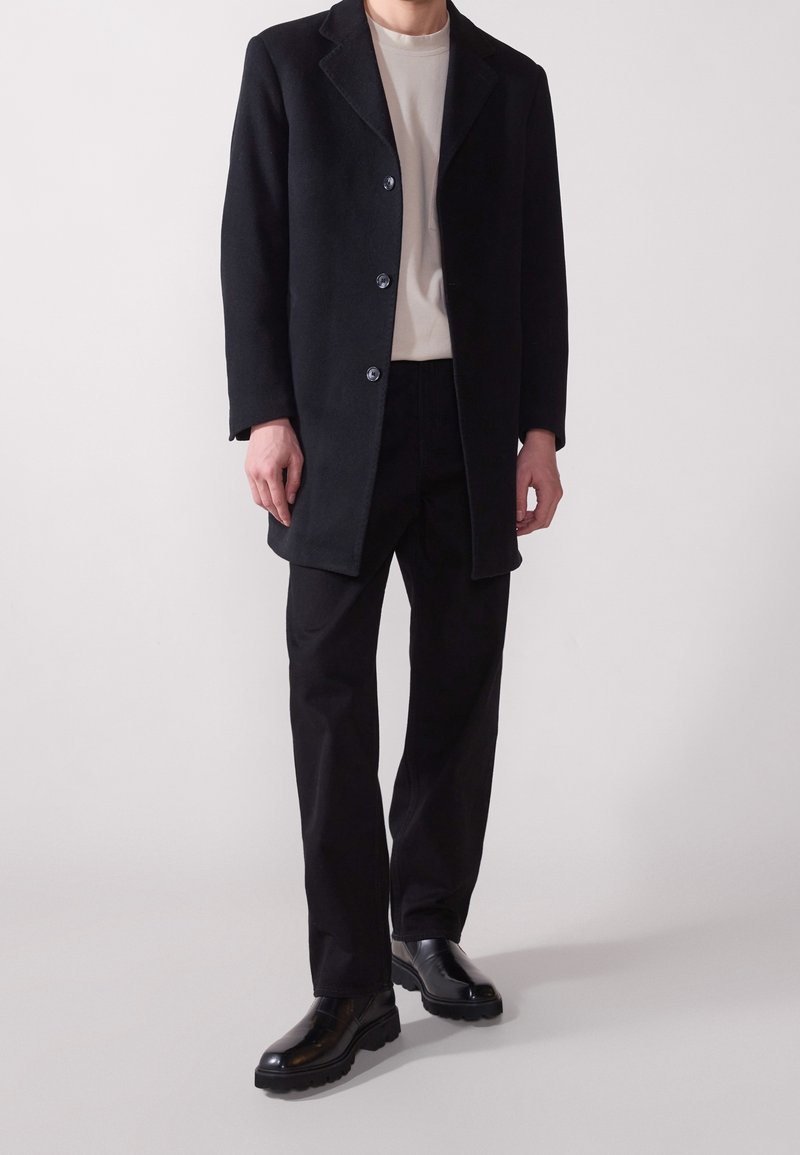 Manteau en laine noire avec col cranté, doté d'une fermeture à boutons. Associé à une chemise beige clair et un pantalon noir, complété par des bottes noires.