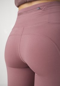 Gros plan sur une personne portant un legging taille haute mauve avec des coutures visibles et une petite poche zippée à la taille.