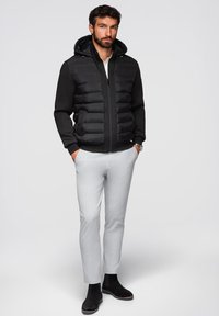 Giacca a vento nera con cappuccio, zip frontale e polsini a coste, abbinata a pantaloni slim-fit grigio chiaro e stivaletti neri.