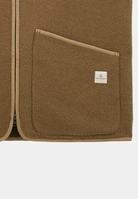 Gilet en polaire marron avec fermeture éclair, doté d'une poche avant et de finitions en tan contrasté, ainsi qu'une étiquette avec la mention "SERGE BLANCO."