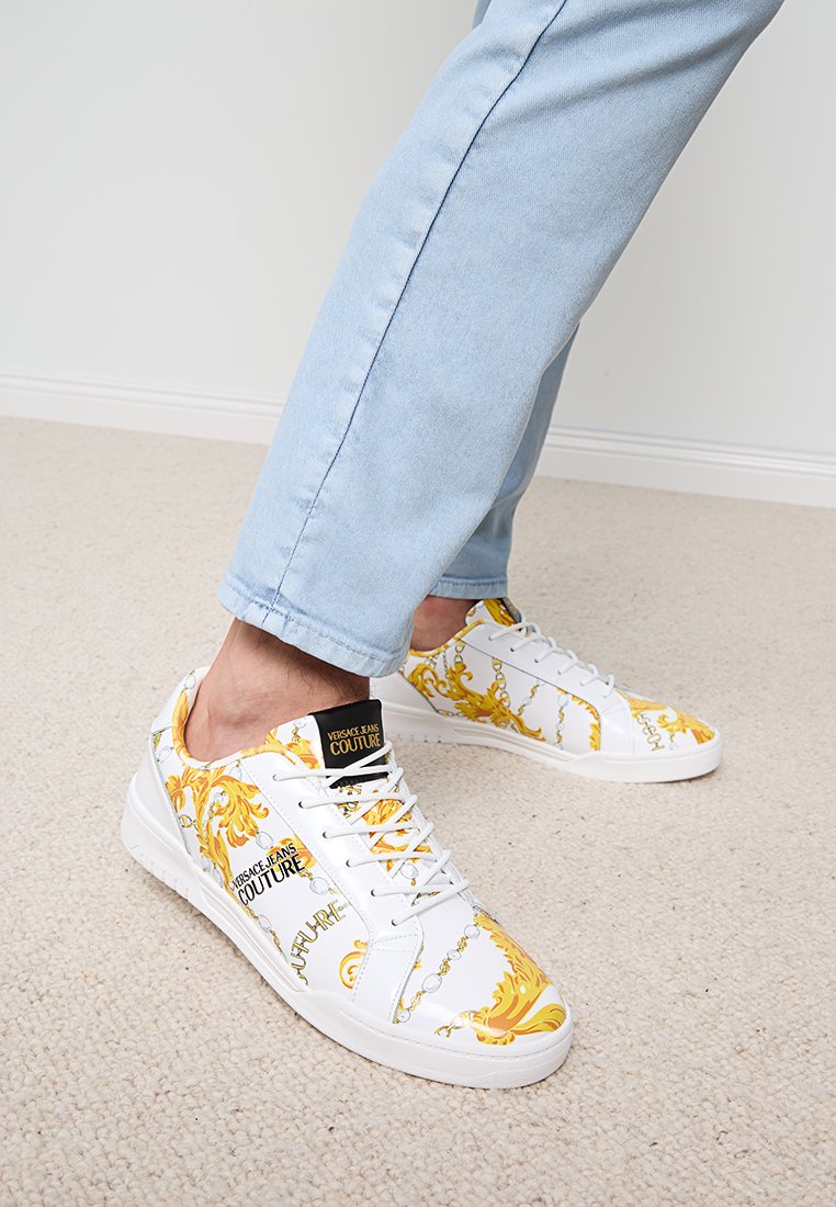 Baskets blanches ornées de motifs baroques jaunes, avec une étiquette noire portant les mots "Versace Jeans Couture". Cuir lisse et semelle plate.