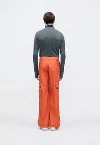 Cilvēks stāv ar muguru, tērpies pelēkā garajās piedurknēs krekls un spilgti oranžas platās kabatas bikses ar baltām kurpēm.