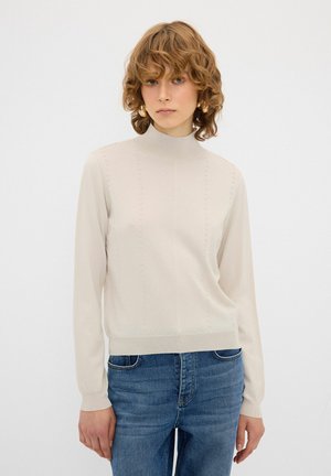 Helles beiges Strickpullover mit hohem Kragen, gerippten Bündchen und einem strukturierten, gepunkteten Muster an der Vorderseite. Kombiniert mit blauen Jeans.