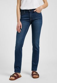 Jeans de denim azul oscuro con un corte ajustado, cinco bolsillos y un ligero estiramiento. Combinados con sandalias marrones y texturizadas que cuentan con una banda decorativa.