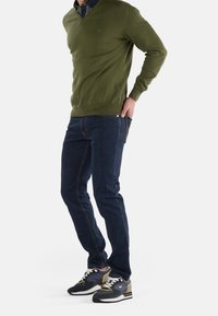 Zielony sweter w serek, ciemnoniebieskie dżinsy i wielokolorowe sneakersy. Sweter ma gładką fakturę; dżinsy mają prosty krój; sneakersy mają sznurówki i akcenty.