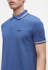 Homme portant un polo bleu avec une patte de boutonnage à trois boutons, un col et des bordures de manches rayés noir et blanc, et "BOSS" brodé sur la poitrine.