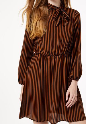 Femme portant une robe à rayures verticales marron et noires avec des manches longues et un nœud noué au niveau du cou, taille cintrée, main détendue le long du corps.