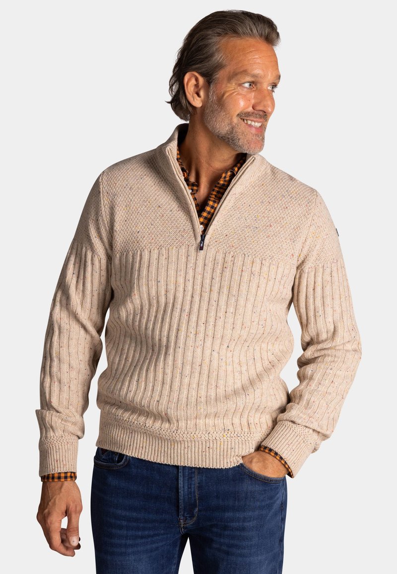 Lichtbeige trui met ribbelstructuur en spikkels. Voorzien van een halve rits kraag en aansluitende manchetten, gecombineerd met donkerblauwe jeans.